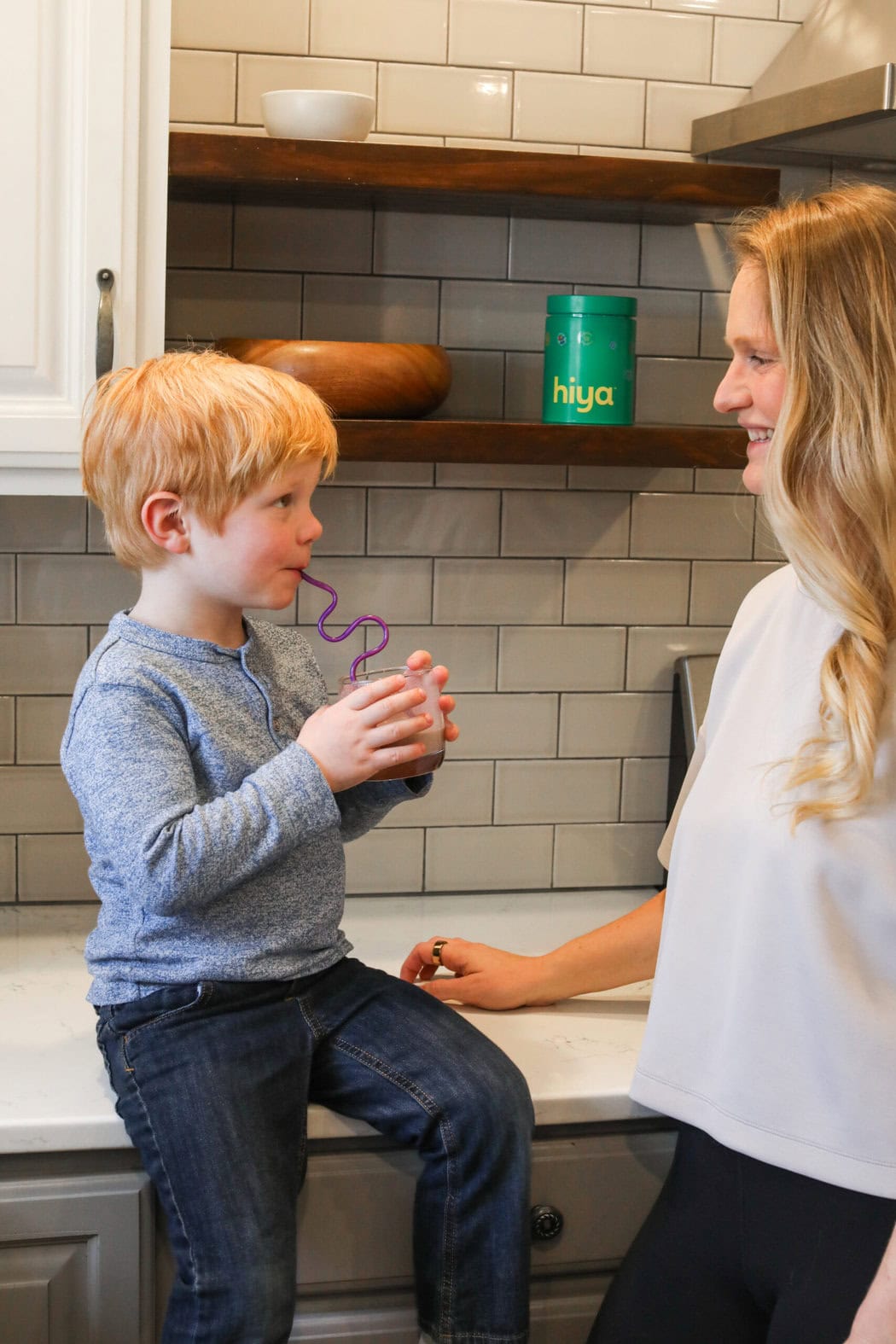 A mother and son in a kitchen smiling at each other while the boy sips a chocolate milk with a curly straw.