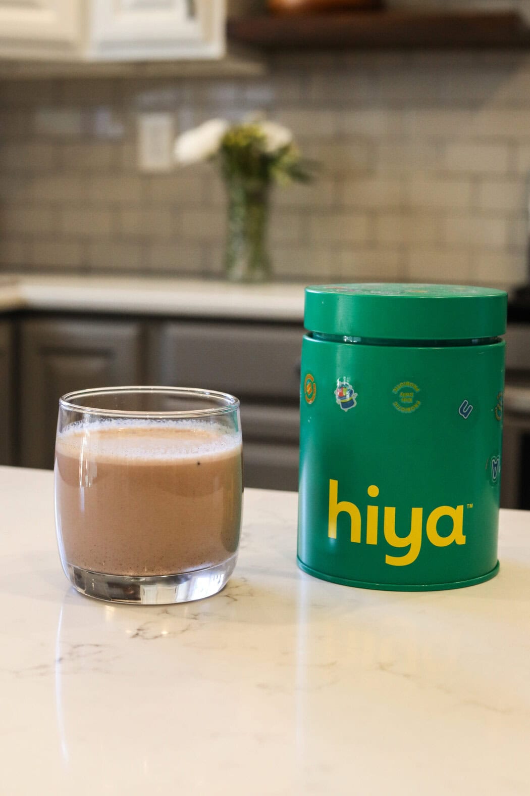 Close up view of a glass of hiya chocolate drink mix next to a canister on a white marble kitchen countertop.