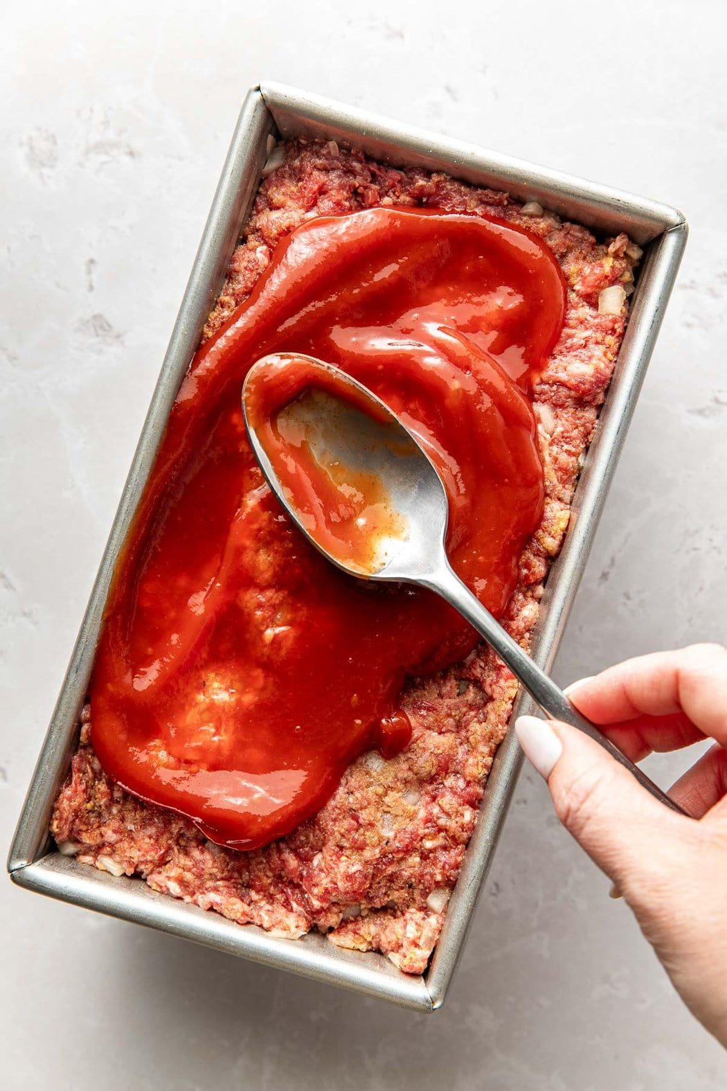 Overhead view of a loaf pan filled with meat loaf mixture being topped with ketchup.