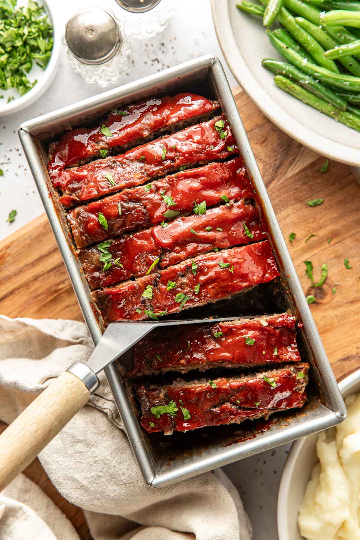 Overhead view of a metal loaf pan filled with freshly baked meatloaf topped with a tomato sauce and fresh herbs.