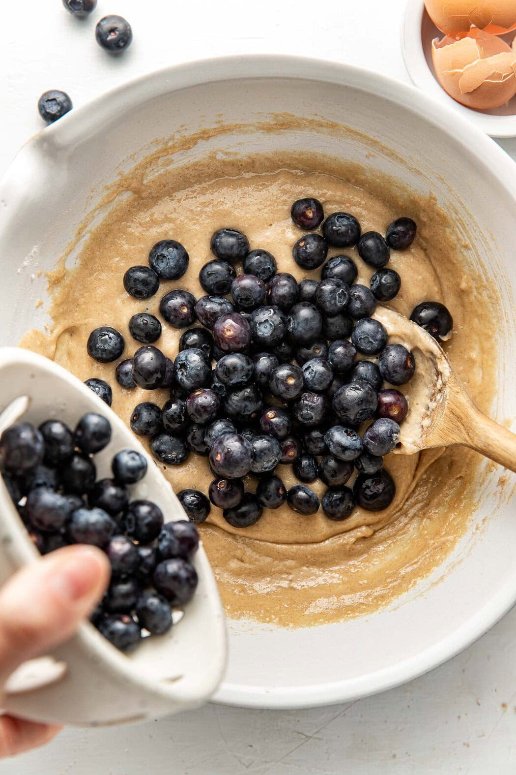 Overhead view of a bowl of coffee cake batter with a hand pouring a bowl of blueberries into the mixture.