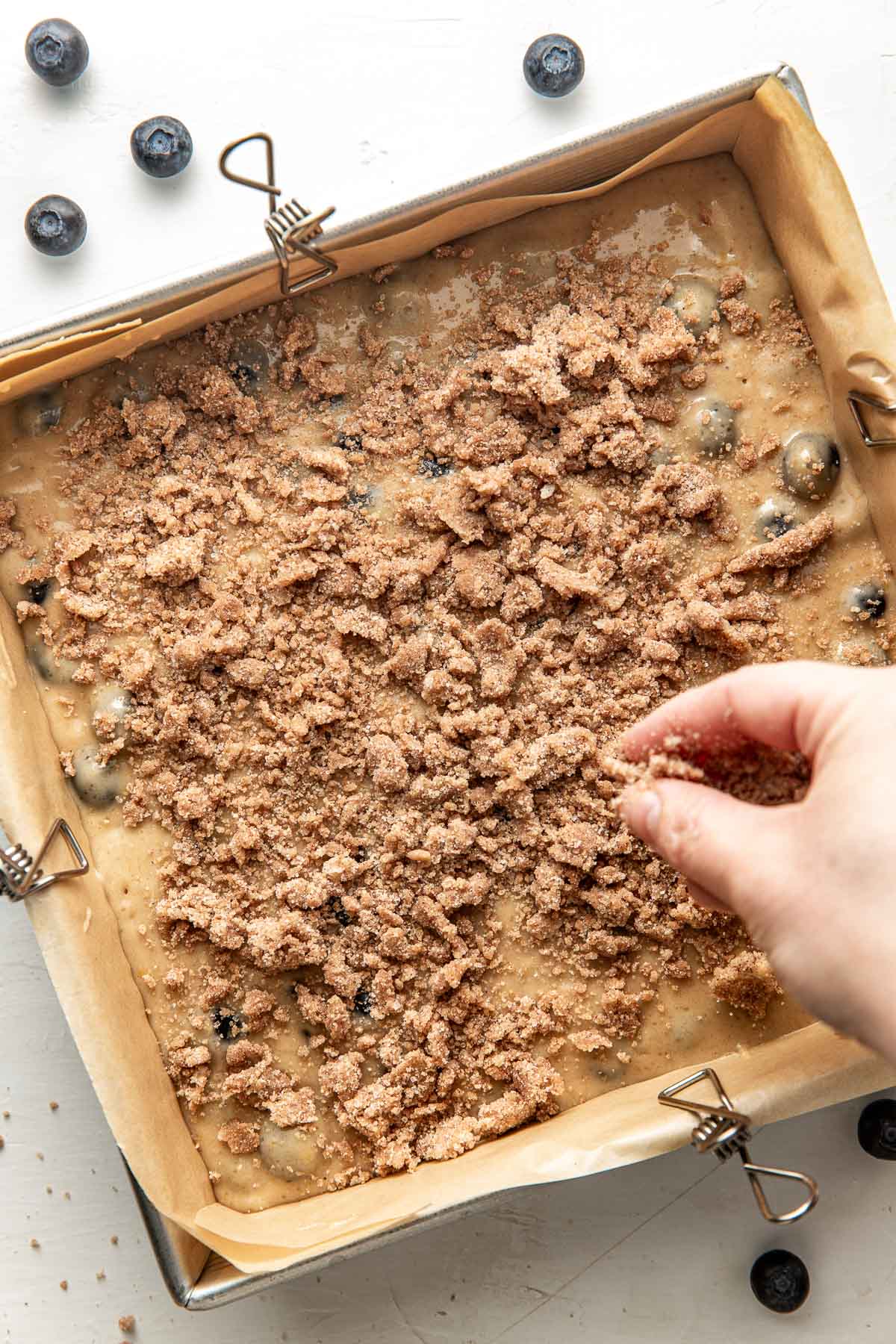 Close up view of a hand sprinkling a crumble topping on to a pan of coffee cake batter. 