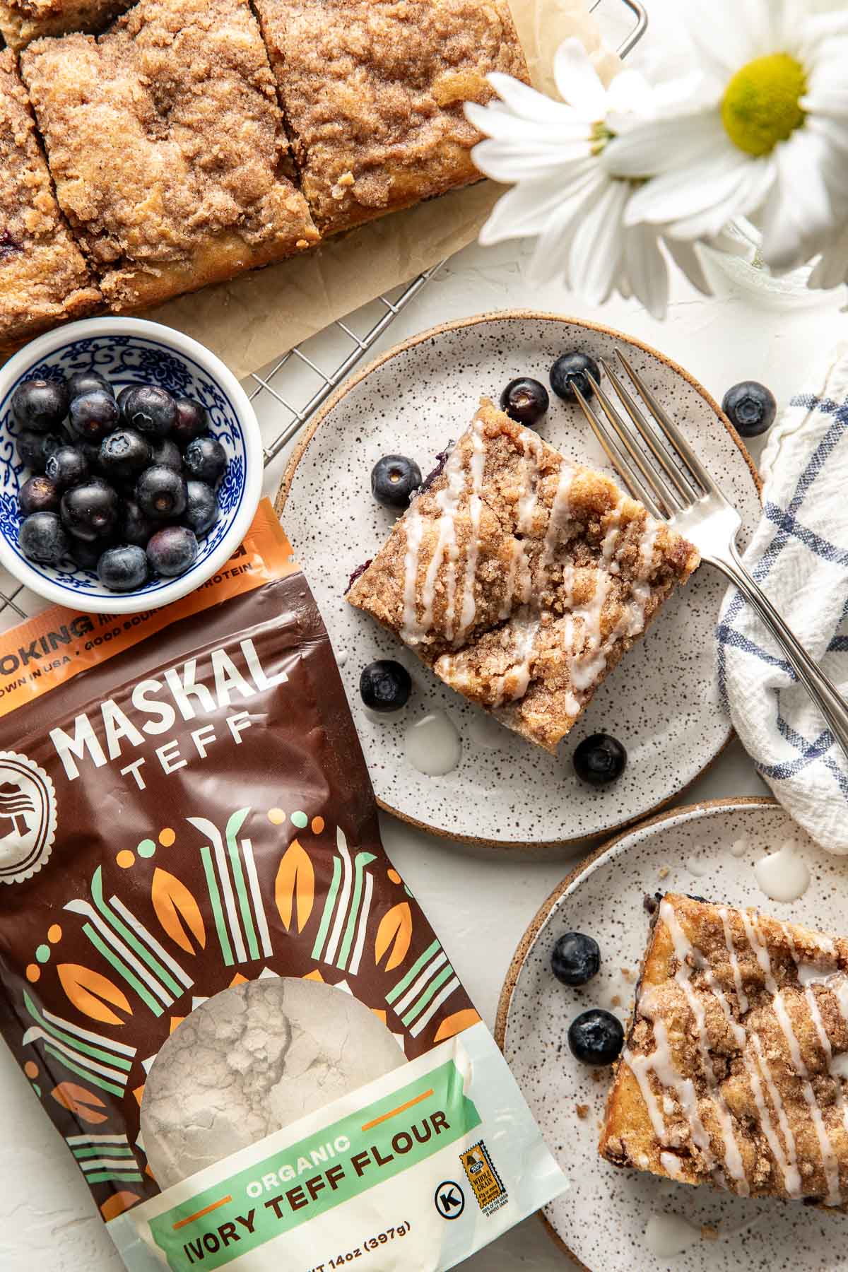 Overhead view of a freshly baked Blueberry Coffee Cake on parchment paper with a small plated piece on the side. 