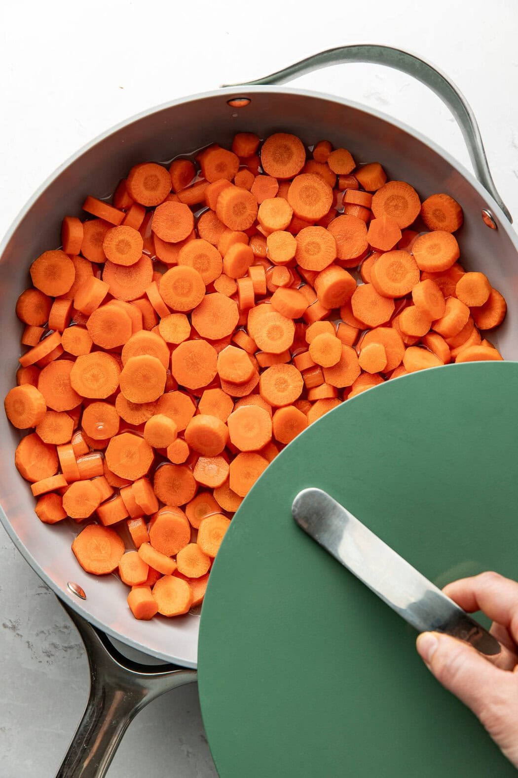 Overhead view of a frying pan filled with sliced carrots and a hand placing the lid on top of the pan.