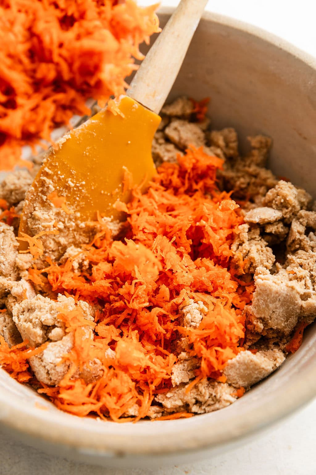 Overhead view of a bowl of carrot cake cookie ingredients including shredded carrots being mixed with a spatula. 