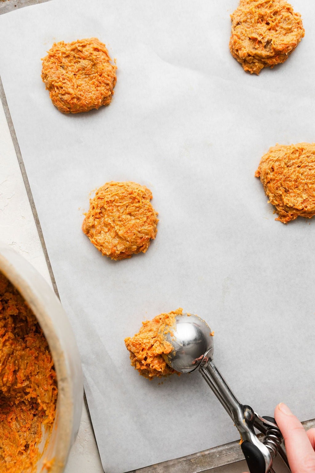 Overhead view of a cookie scoop placing carrot cake cookie dough on a parchment lined sheet pan. 