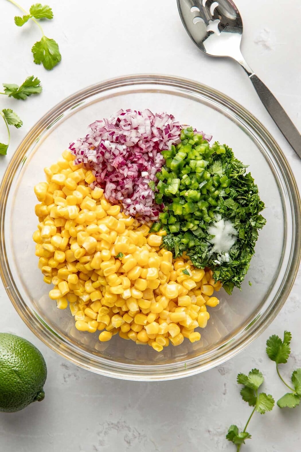 Overhead view of a glass bowl filled with corn, red onions, peppers, and fresh cilantro ready for mixing. 
