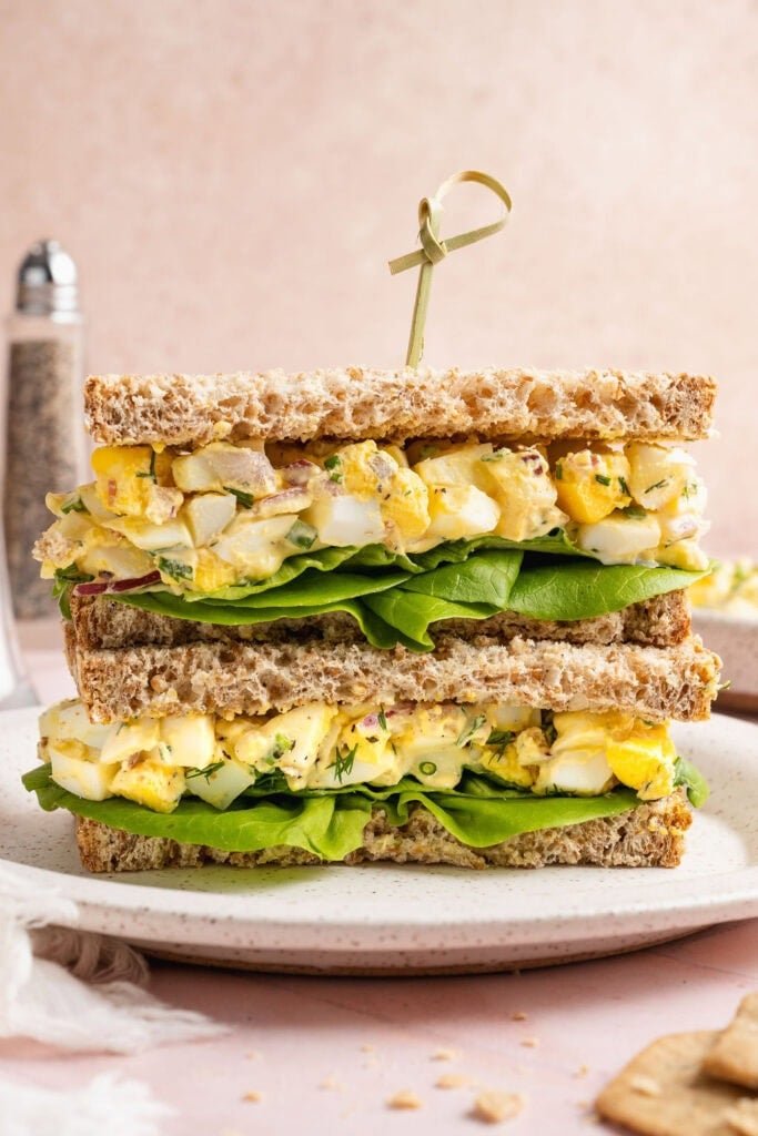 Close up view of an egg salad sandwich halved and stacked showing the layers of bread, lettuce, and egg salad.