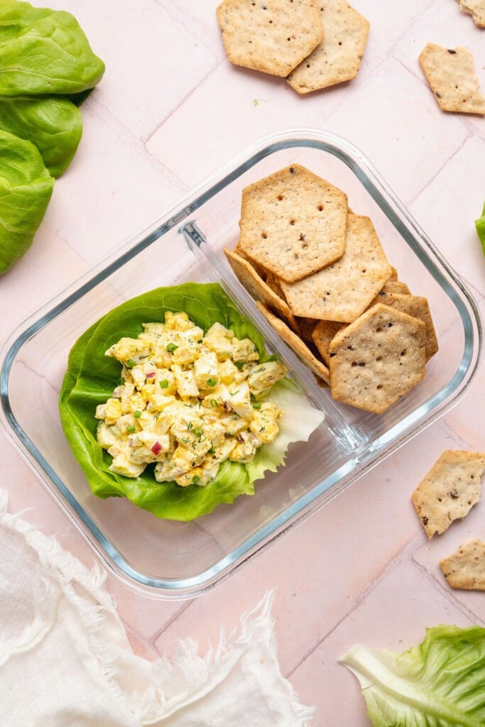 Overhead view of a glass meal prep container with crackers and a lettuce leaf topped with egg salad.