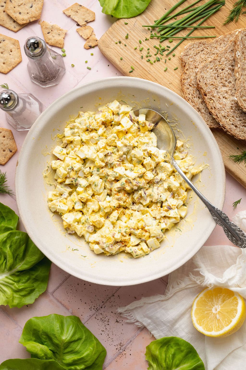 Overhead view of a bowl of egg salad topped with fresh dill and chives surrounded by crackers. 