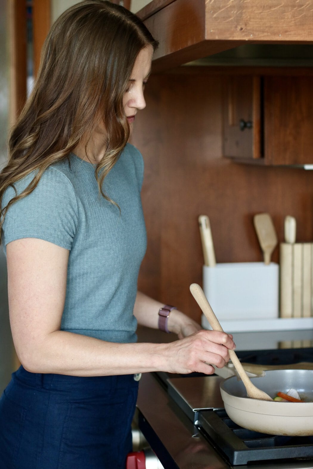 Woman sautéing vegetables in a pan on a stove in a kitchen.