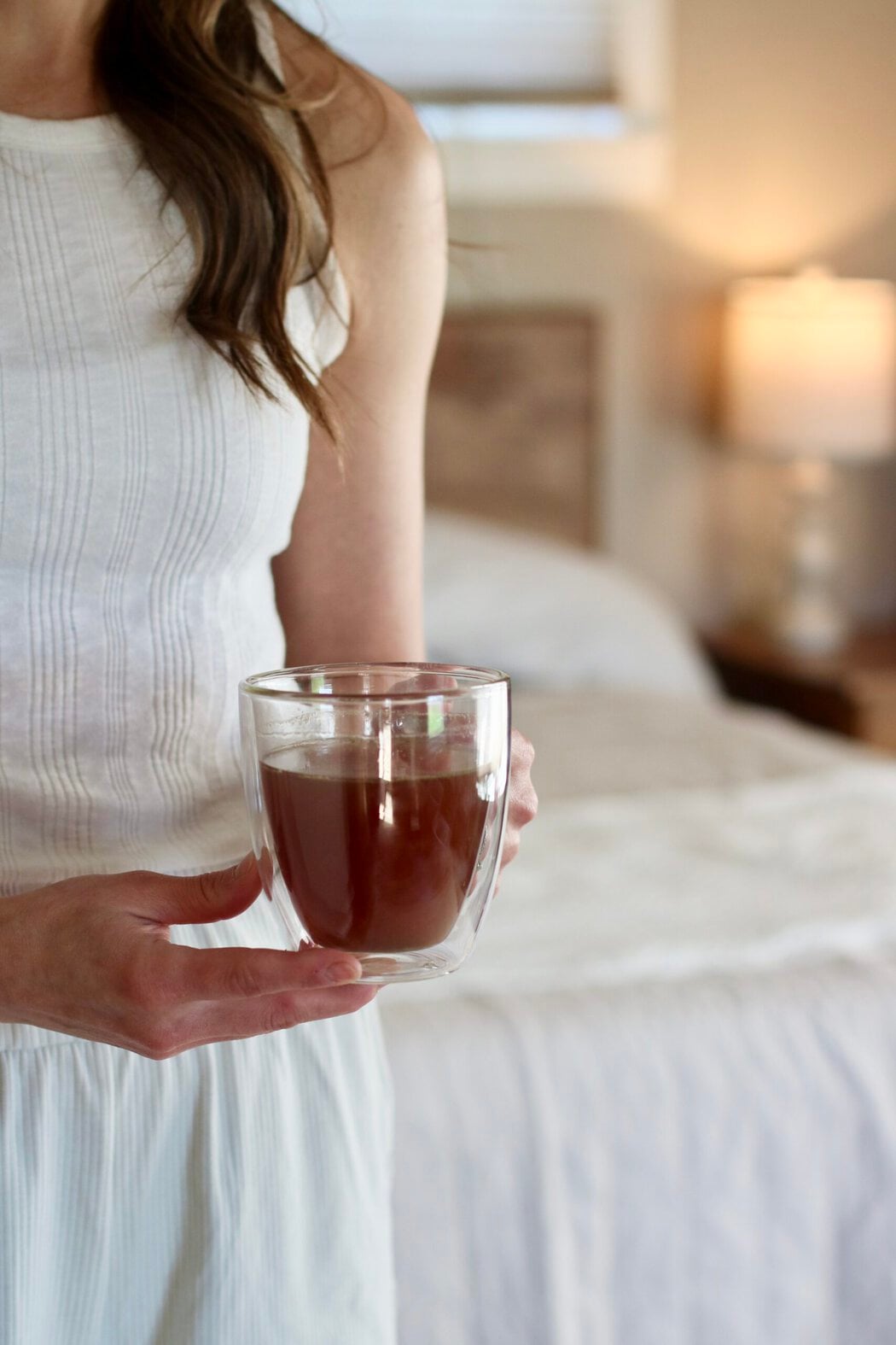 Close up view of a woman in pajamas standing in front of a bed holding a glass mug of nighttime tea.