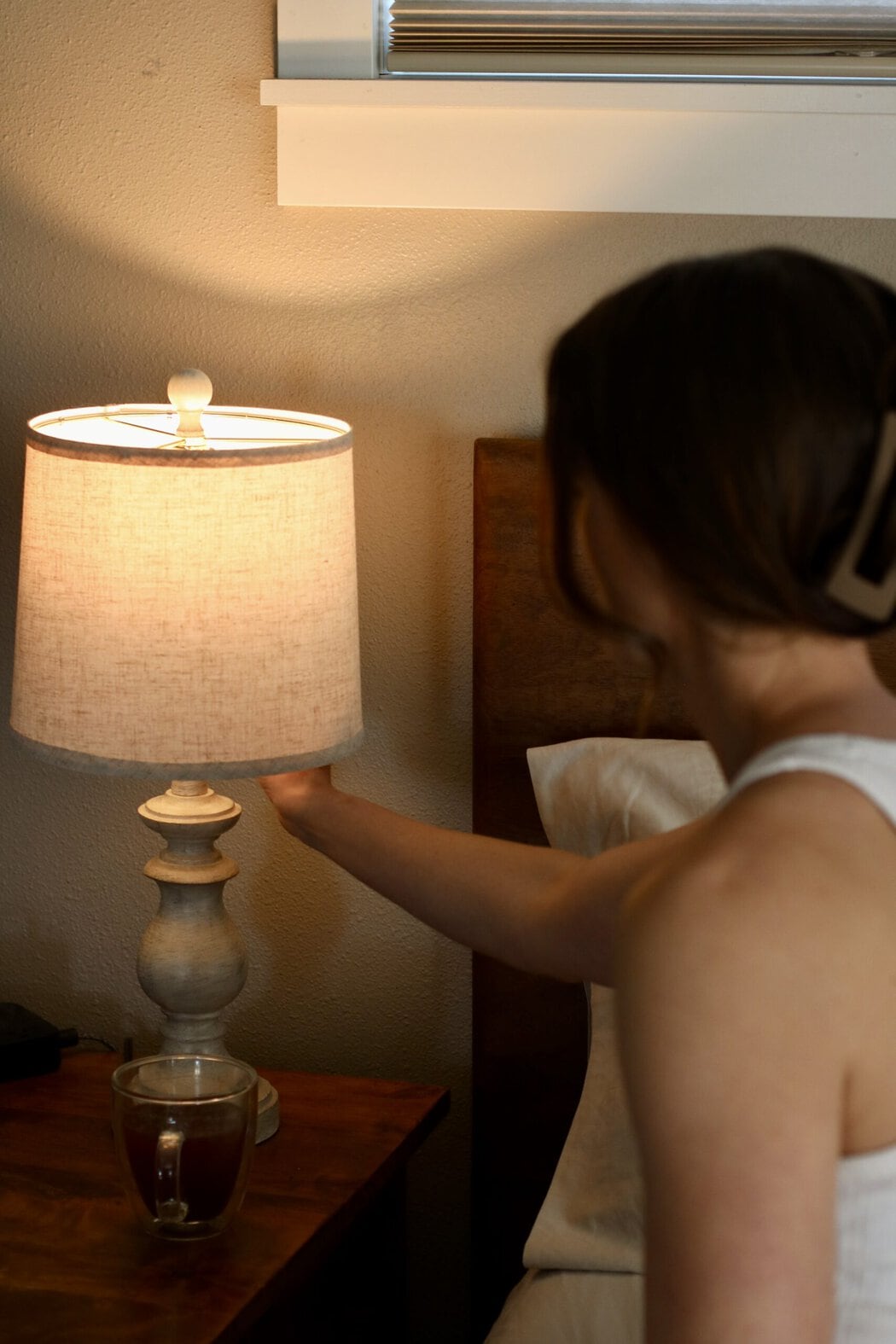 A woman sitting on the edge of a bed reaching over to turn off a bedside lamp.