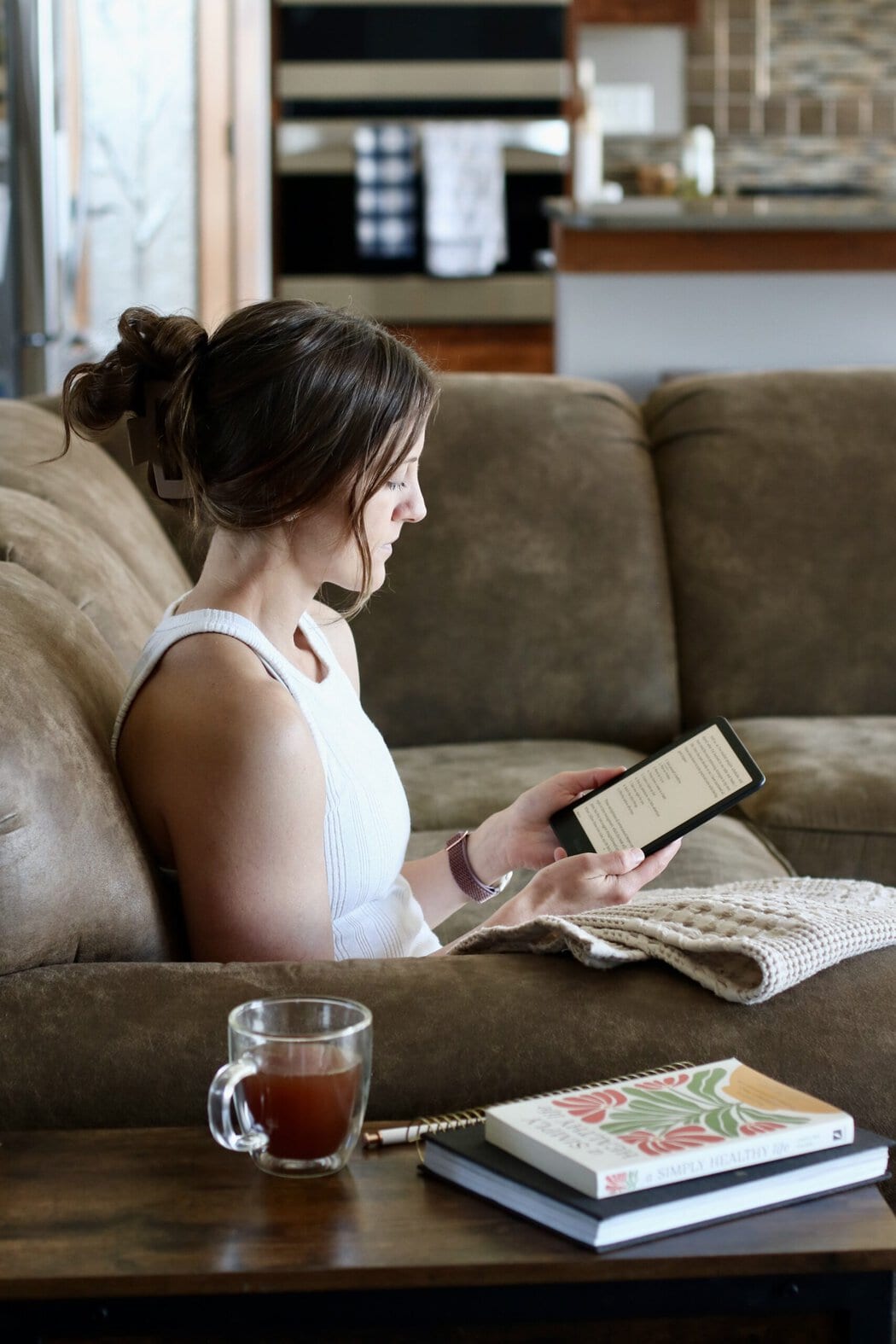 Profile view of a woman sitting on her couch with a blanket on her lap reading from a kindle book.
