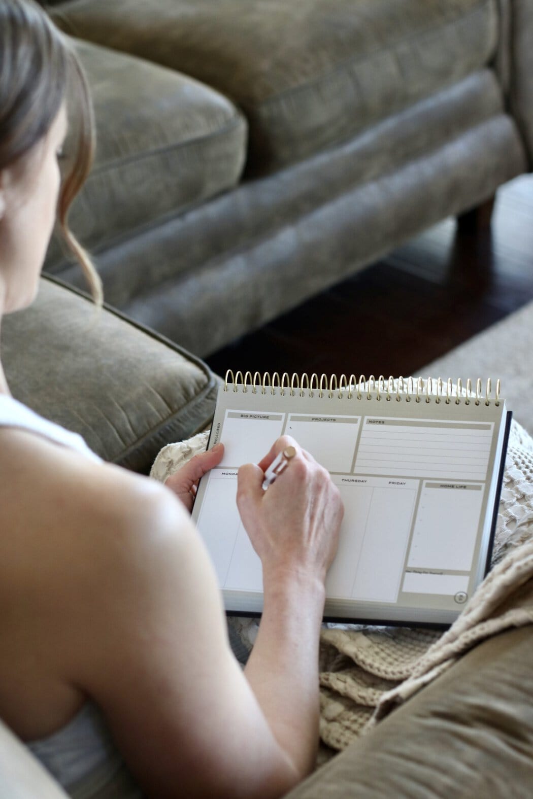 Overhead view of a woman writing out her schedule on a calendar book while sitting on a couch with a blanket on her lap.
