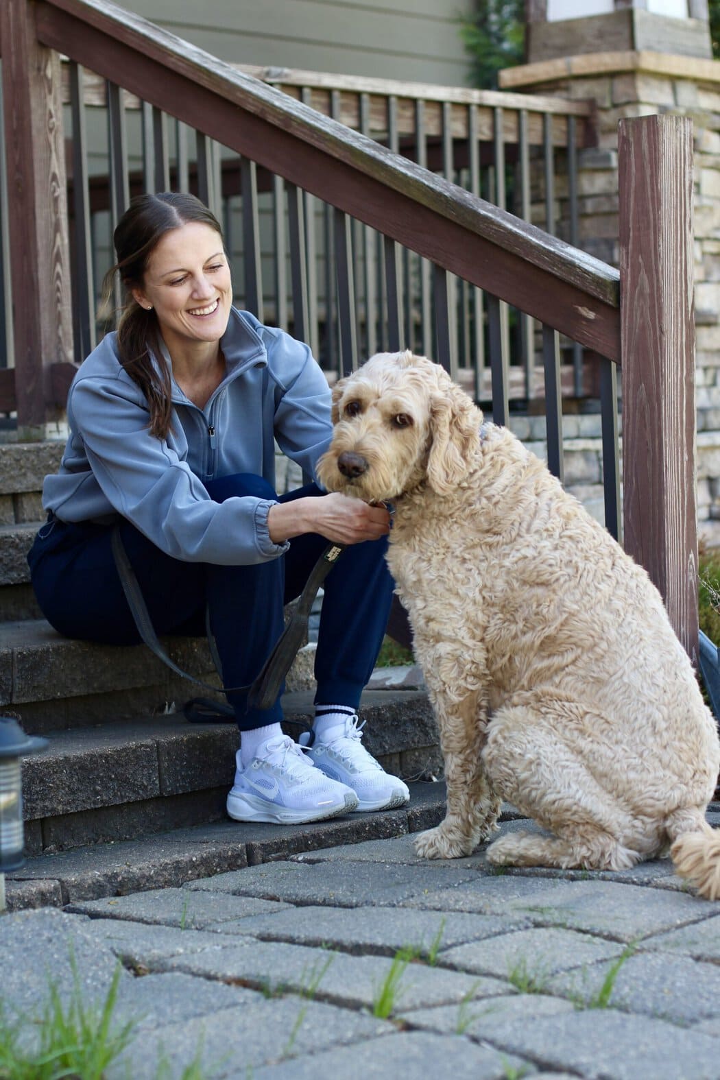 A woman sitting on her front steps leashing up her golden doodle dog for a walk.