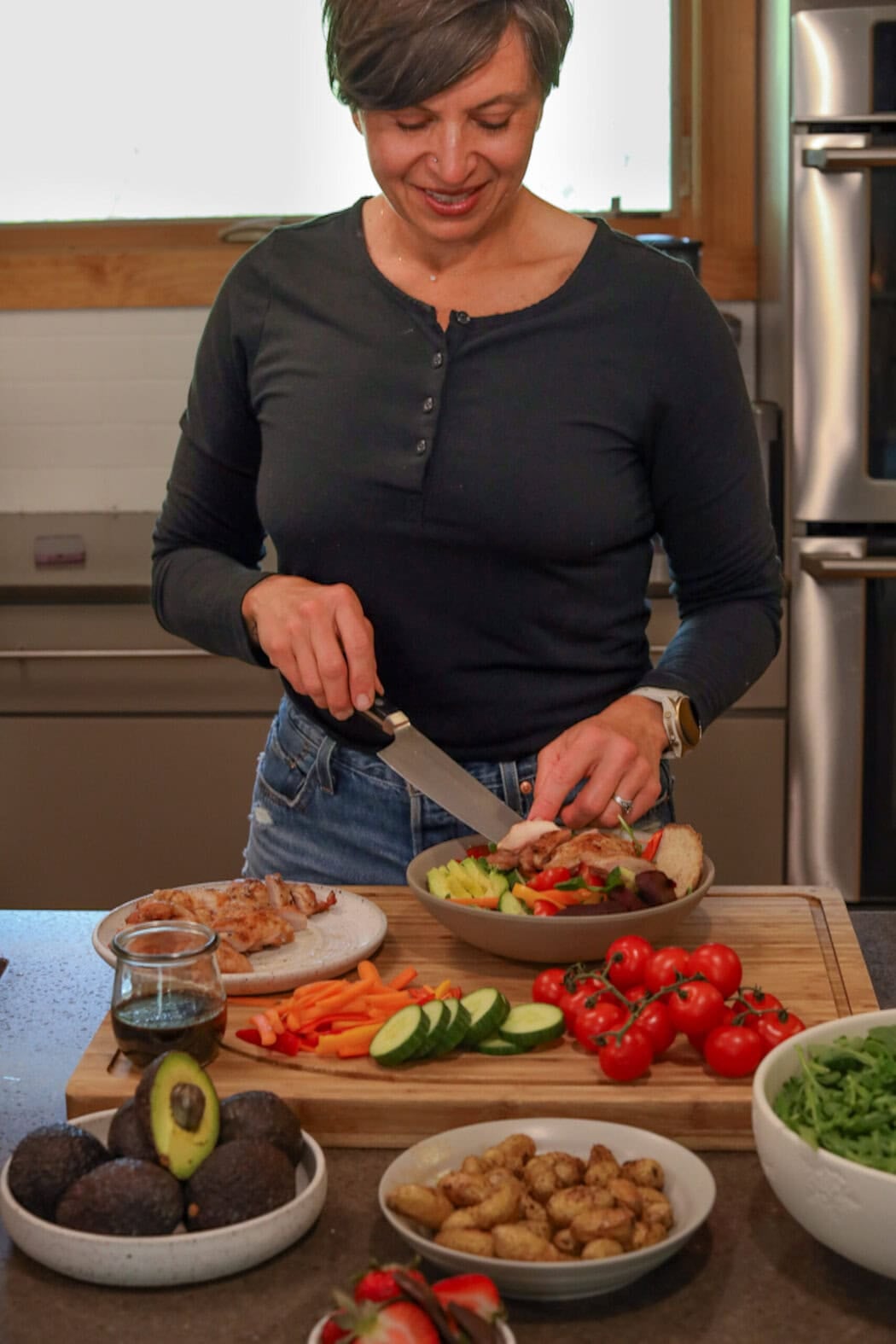 A woman standing at a kitchen countertop slicing and preparing a salad in a bowl. 