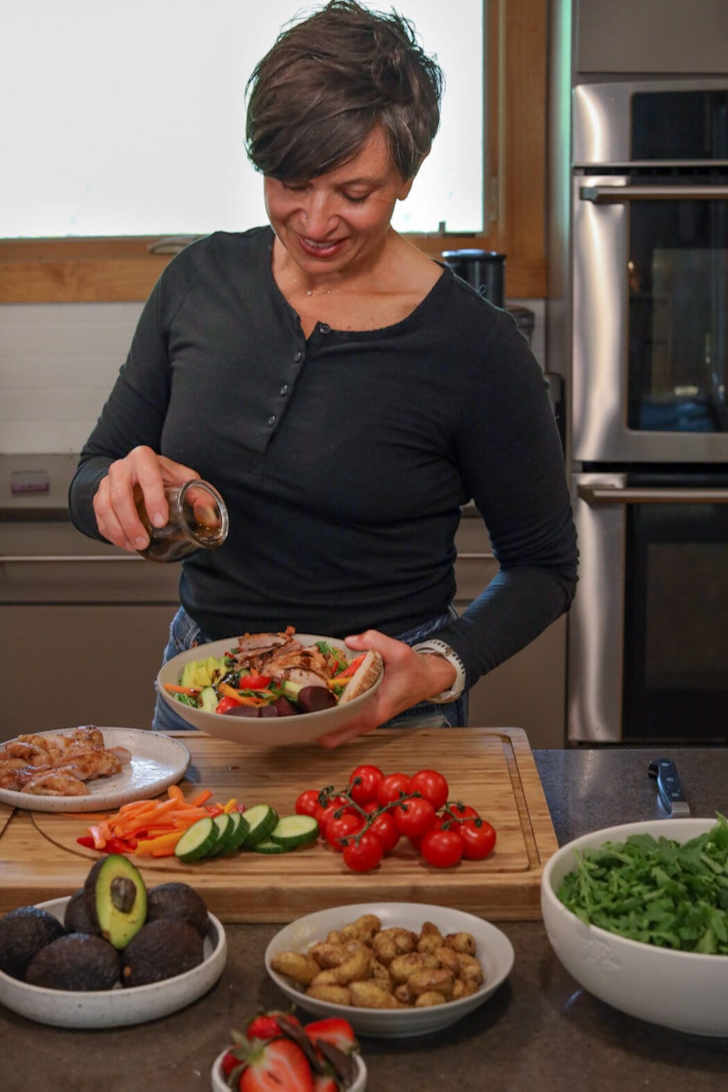 A woman in a kitchen pouring dressing over a bowl of salad by a countertop filled with fresh vegetables. 