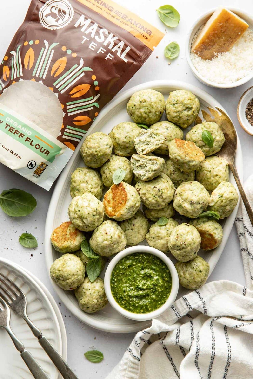 Overhead view of an oval platter of Pesto Meatballs topped with fresh basil and garnished with a small dish of pesto. 