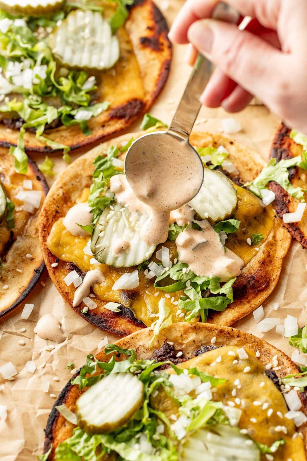 Overhead view of Smash Burger Tacos on parchment paper being drizzled with special sauce and topped with pickles. 