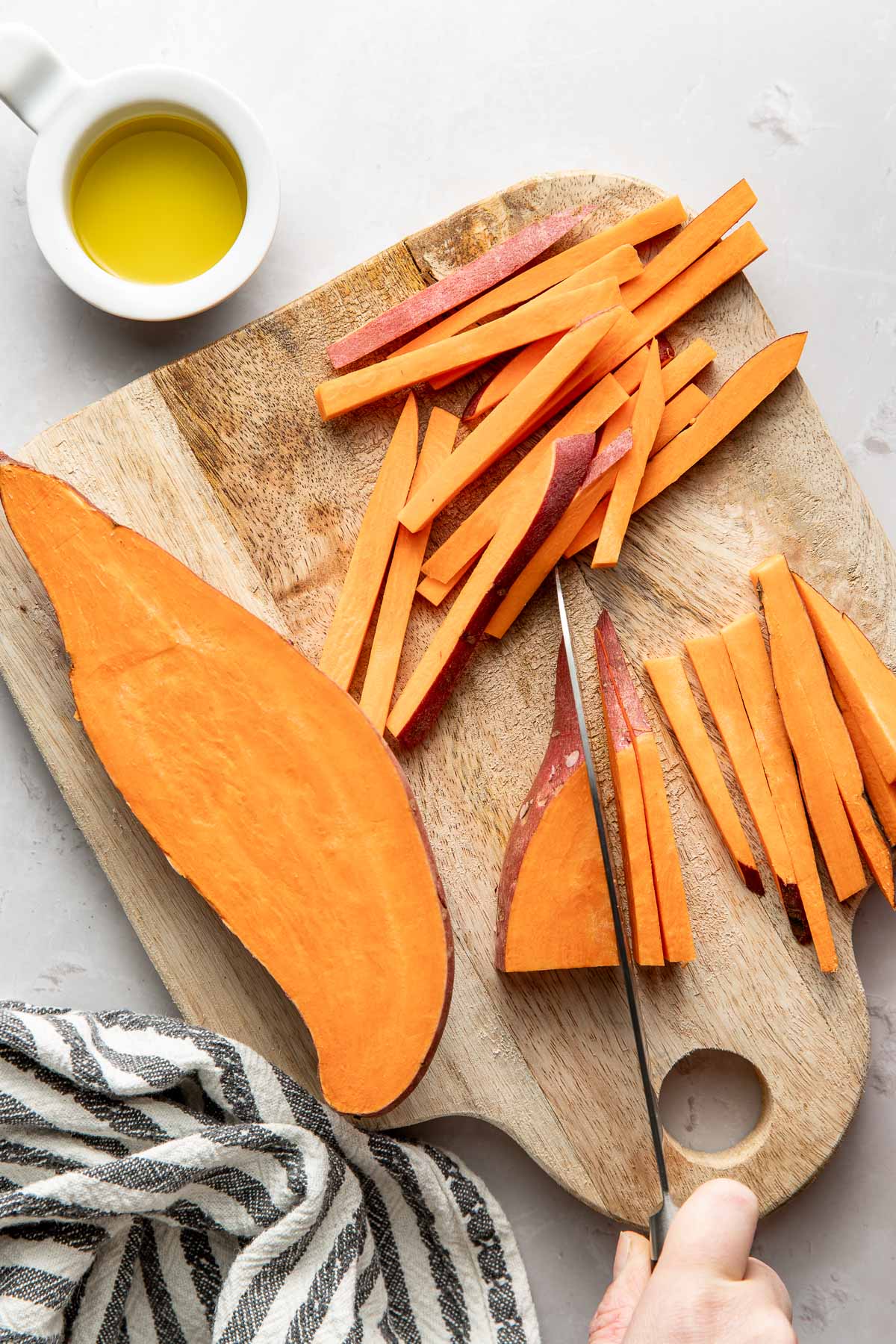 Overhead view of a cutting board with a hand holding a knife slicing a sweet potato into strips for fries.