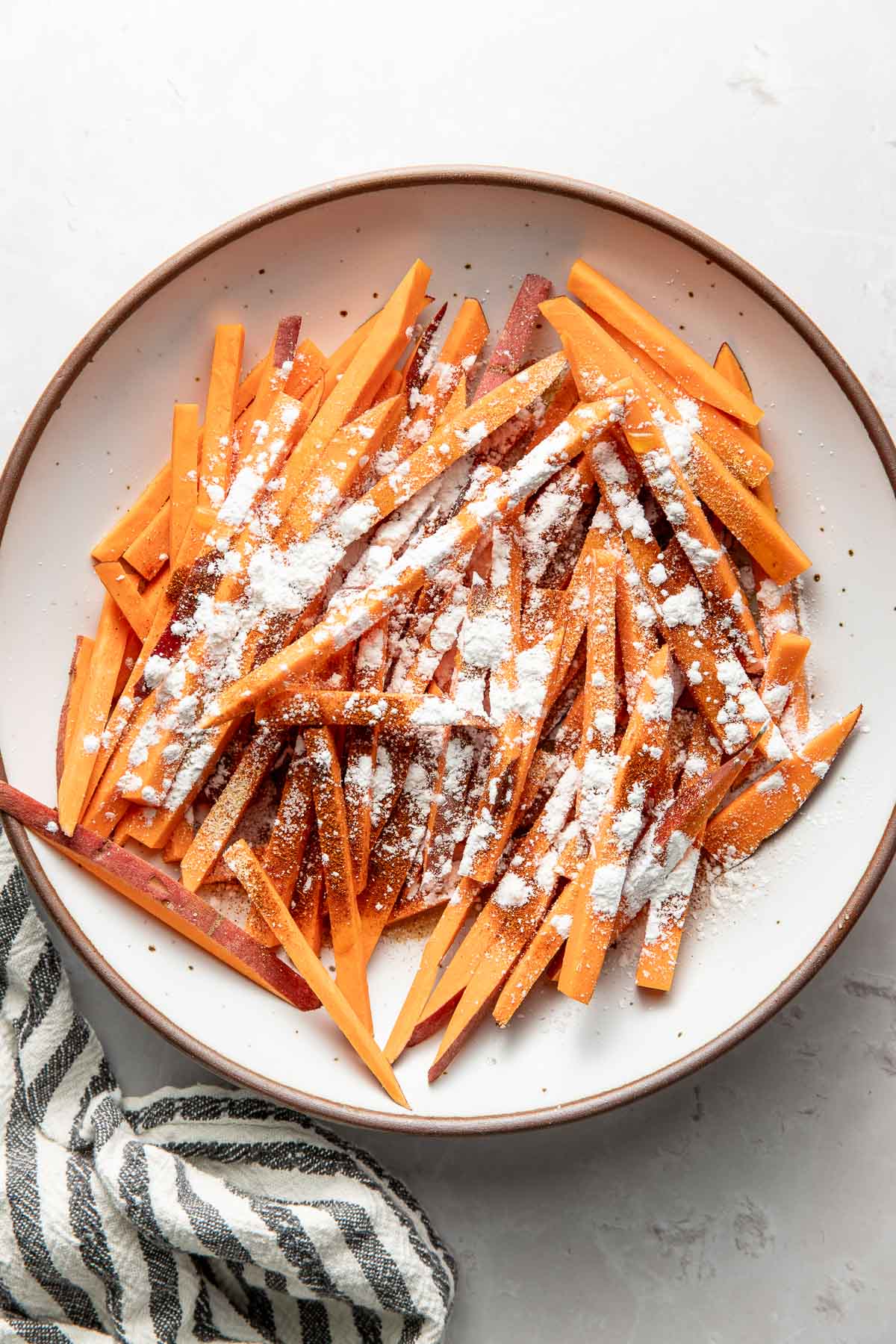 Overhead view of a plate filled with sliced sweet potatoes topped with seasonings and corn starch.