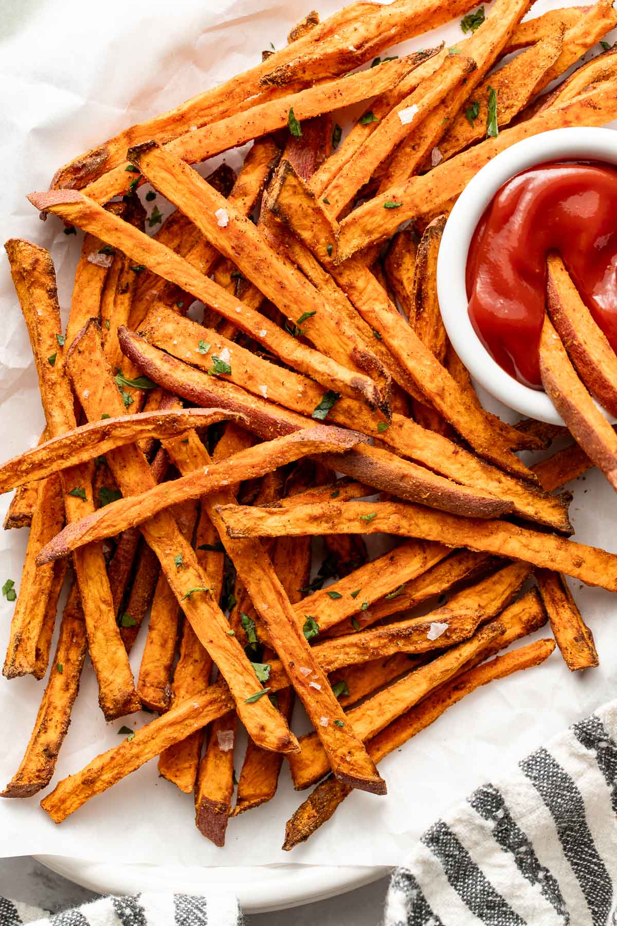 Close up view of Air Fryer Sweet Potato Fries arranged around a small bowl of ketchup topped with flakey sea salt and fresh herbs.