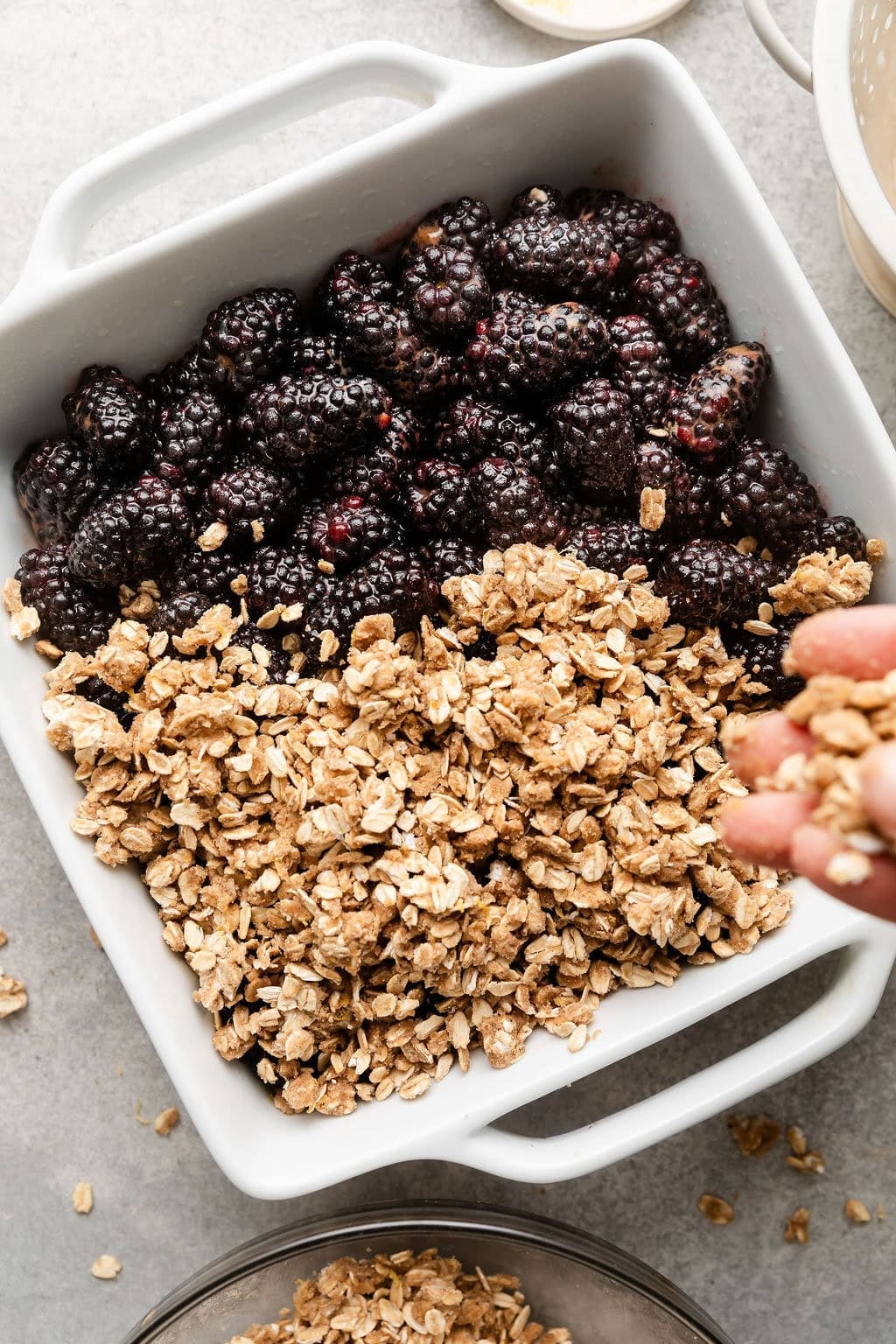 Overhead view of a baking dish filled with fresh blackberries and a hand covering them with an oat mixture on top.