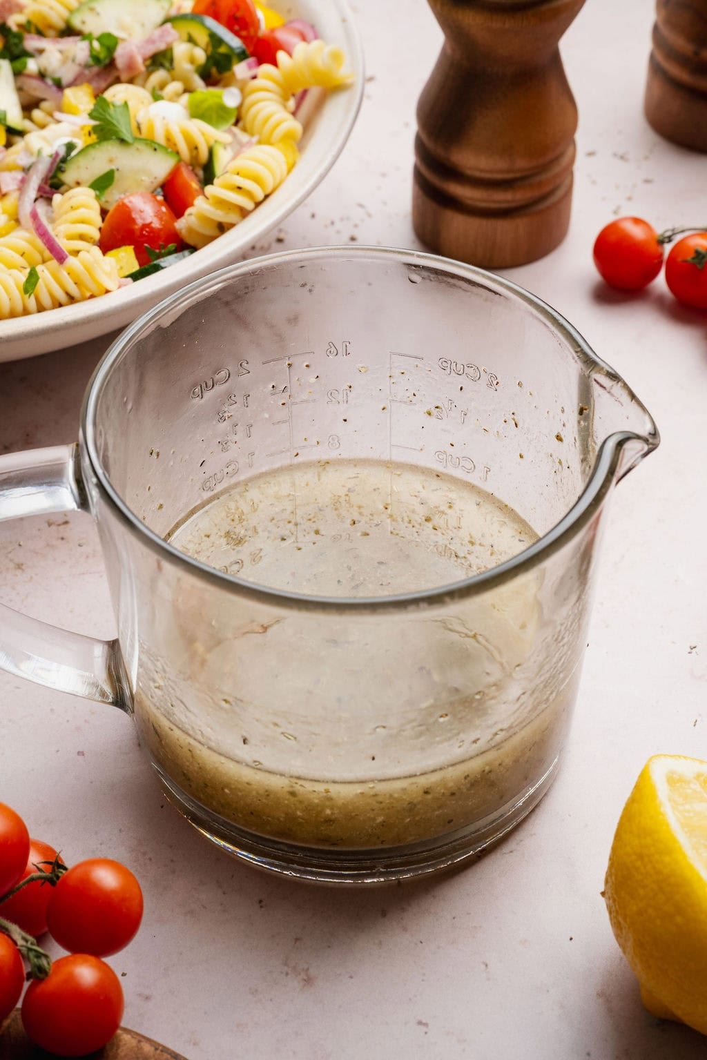 Close up view of a measuring cup filled with Italian Dressing next to a bowl of pasta salad.