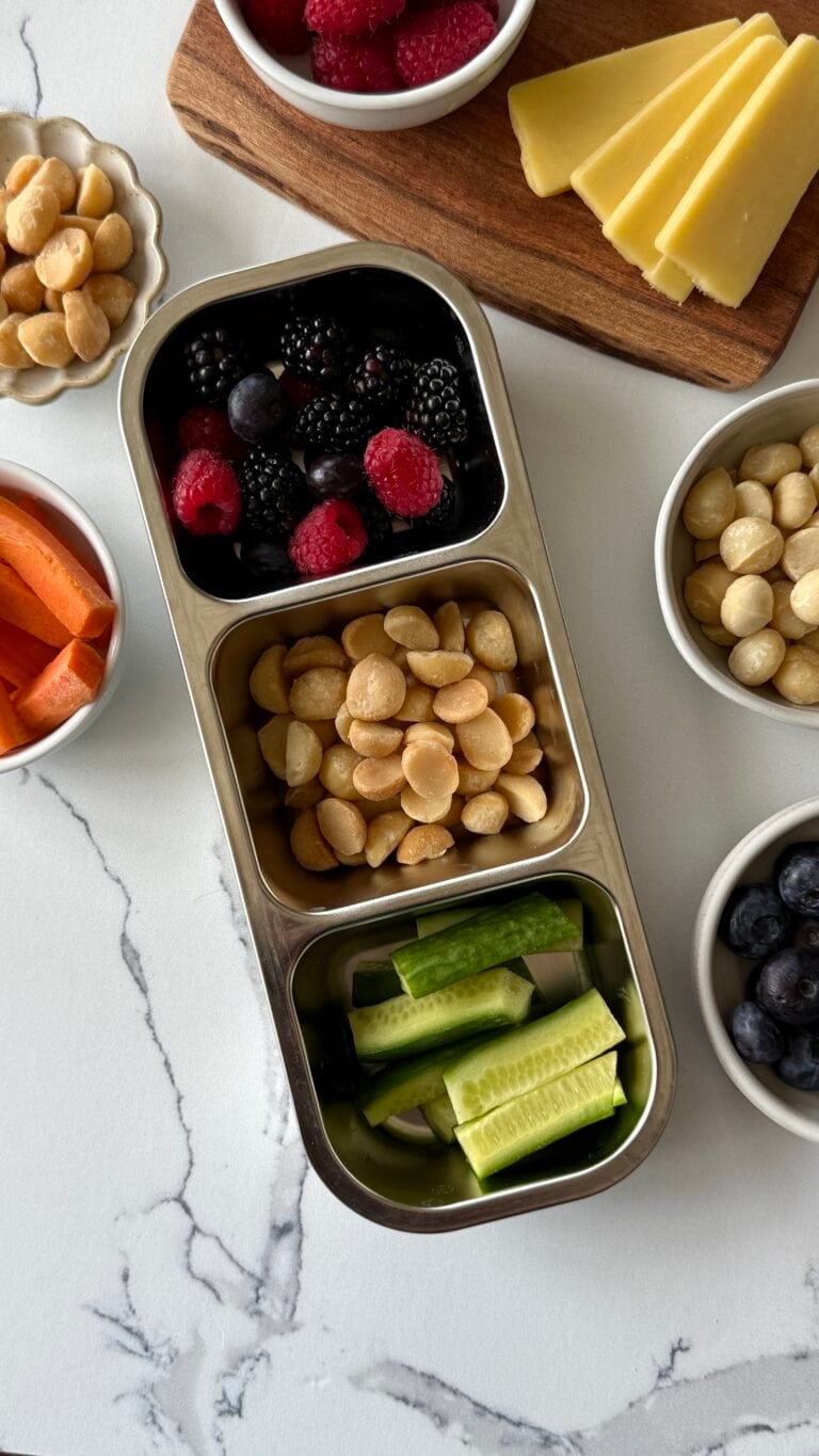 Overhead view of a stainless steel 3-compartment container filled with fresh berries, sliced cucumbers, and macadamia nuts. 