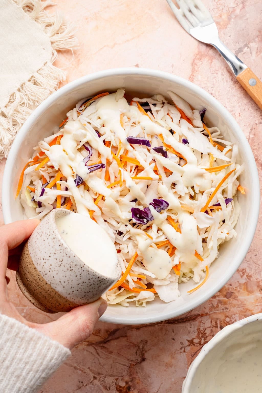 Overhead view of a bowl of cabbage mix with a hand pouring dressing over the top for coleslaw