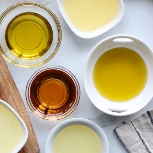 Overhead view of a variety of cooking oils in different sized bowls on a white marble countertop.