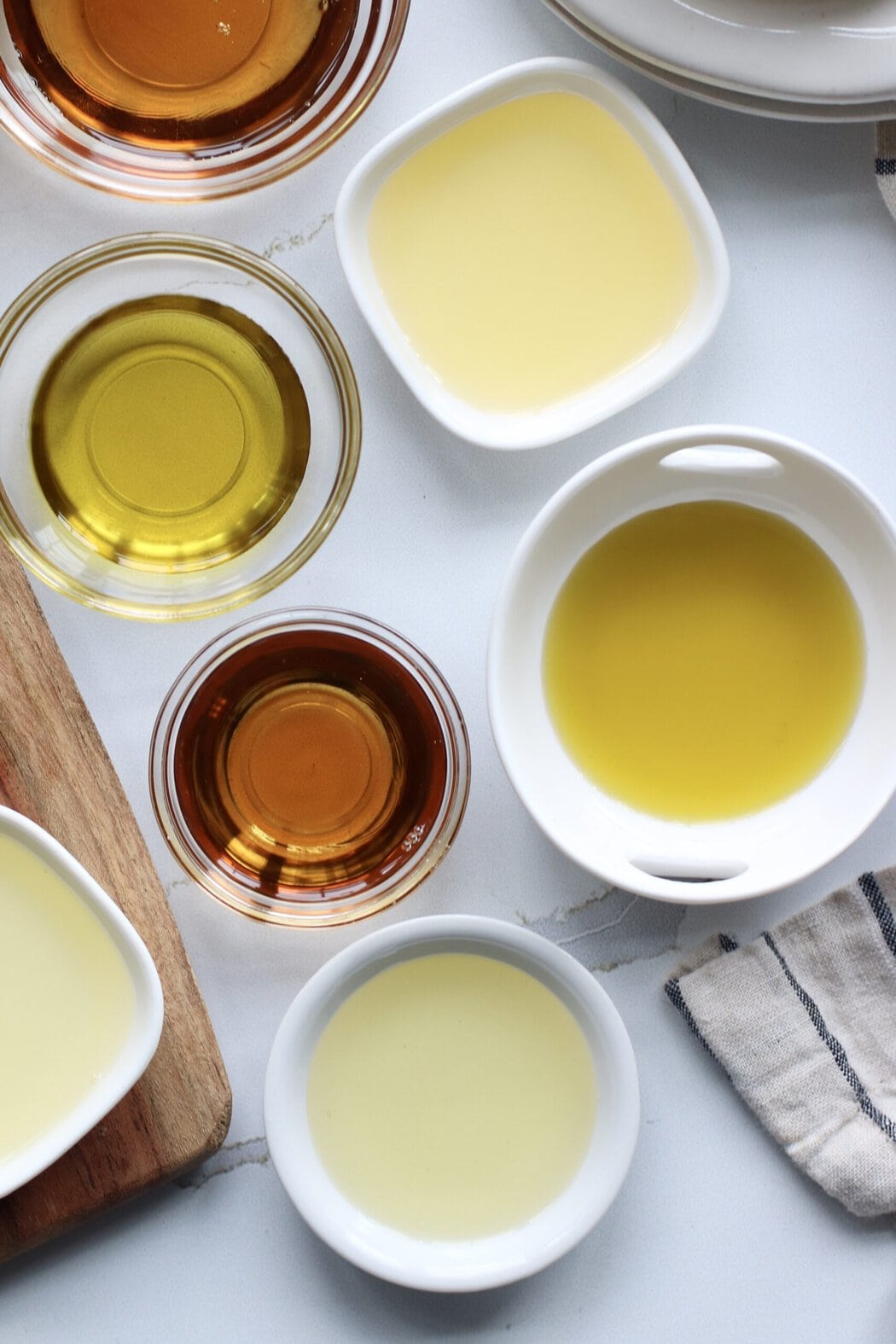 Overhead view of a variety of different colored cooking oils in different sized bowls on a white marble countertop.