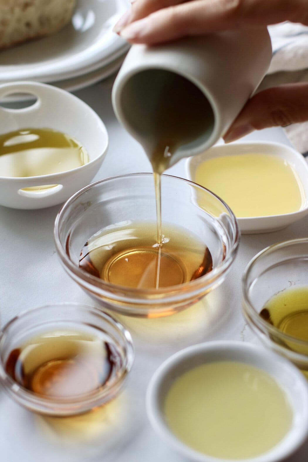 Close up view of a hand pouring cooking oil into a small glass bowl on a white marble countertop.