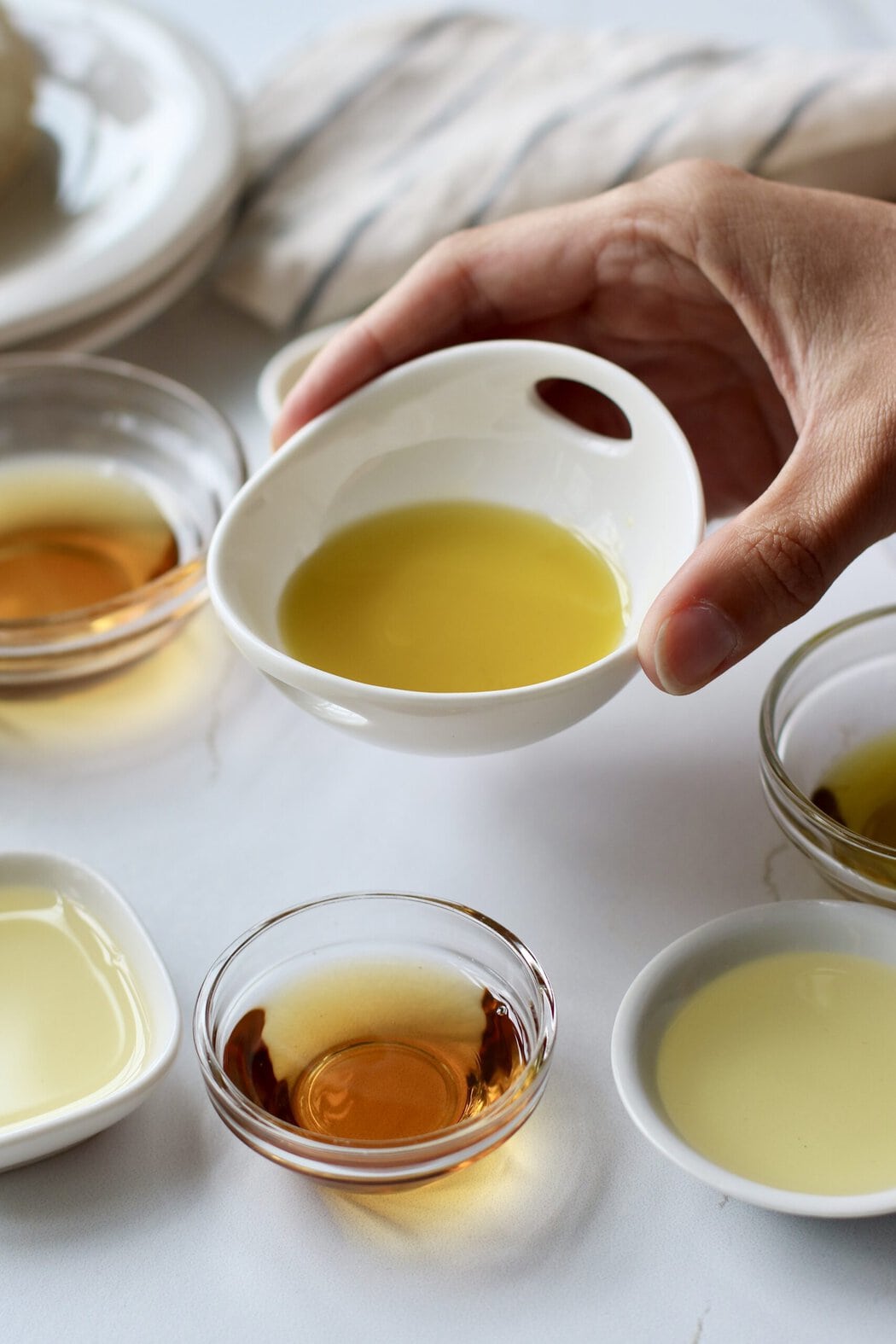 Close up view of a hand holding a small bowl of a cooking oil over a marble white countertop.