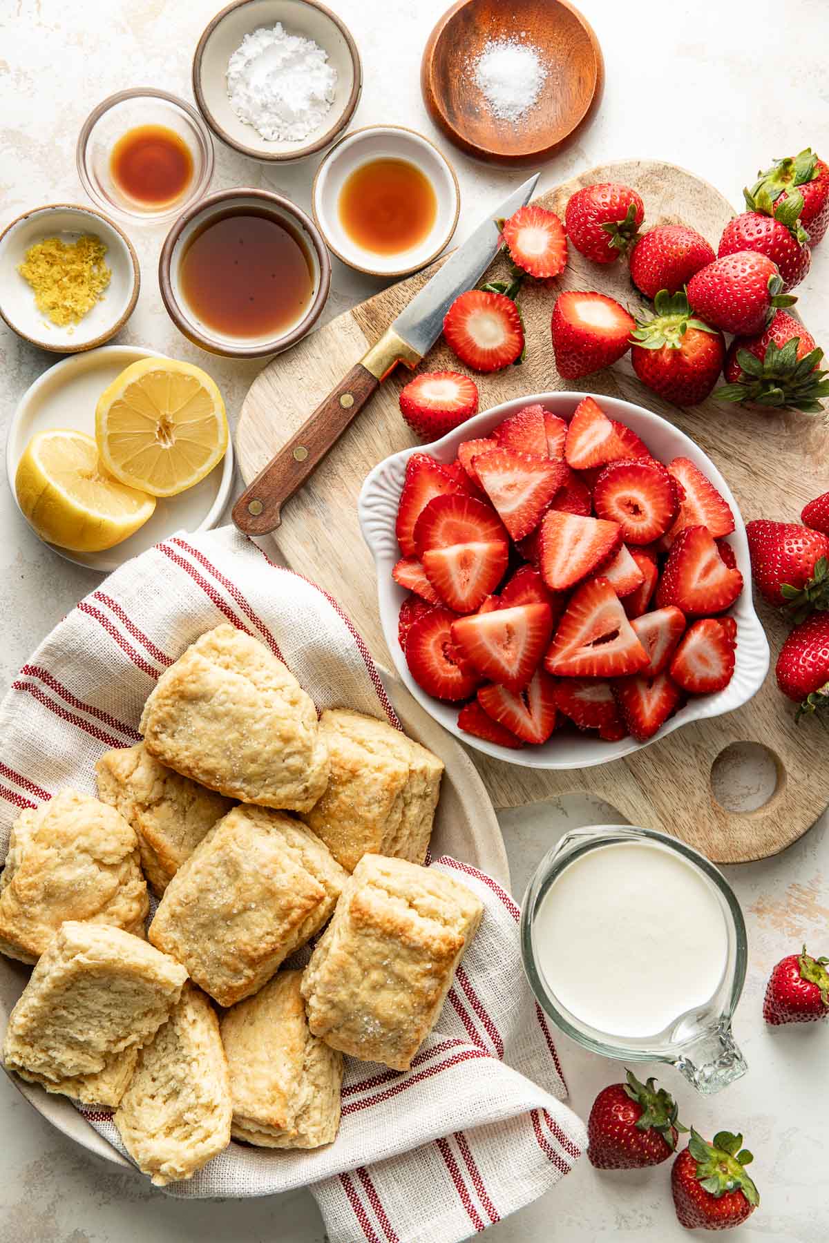 Overhead view of a variety of ingredients for Strawberry Shortcake in different sized bowls and strawberries on a wooden cutting board.