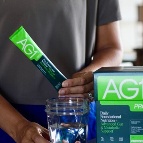 A woman holding an AG1 Pro packet and a glass of water, a box of AG1 Pro packets next to her.