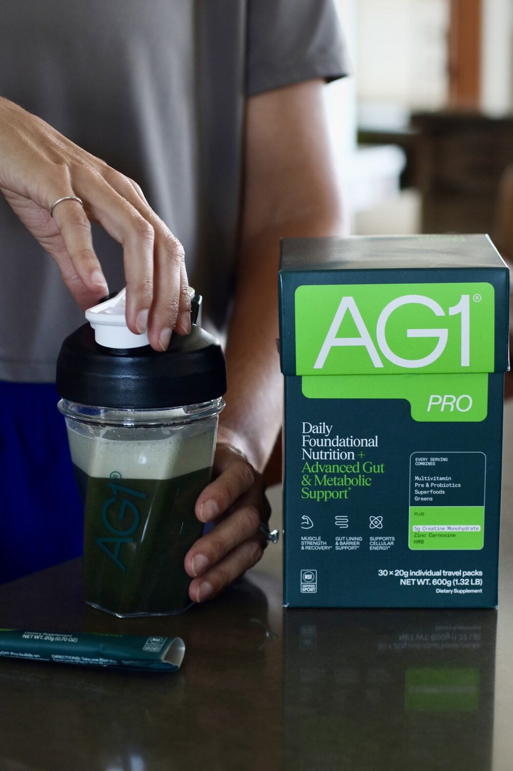 A woman screwing the lid on an AG1 shaker bottle filled with water and a greens packet, a box of AG1 Pro packets next to her. 