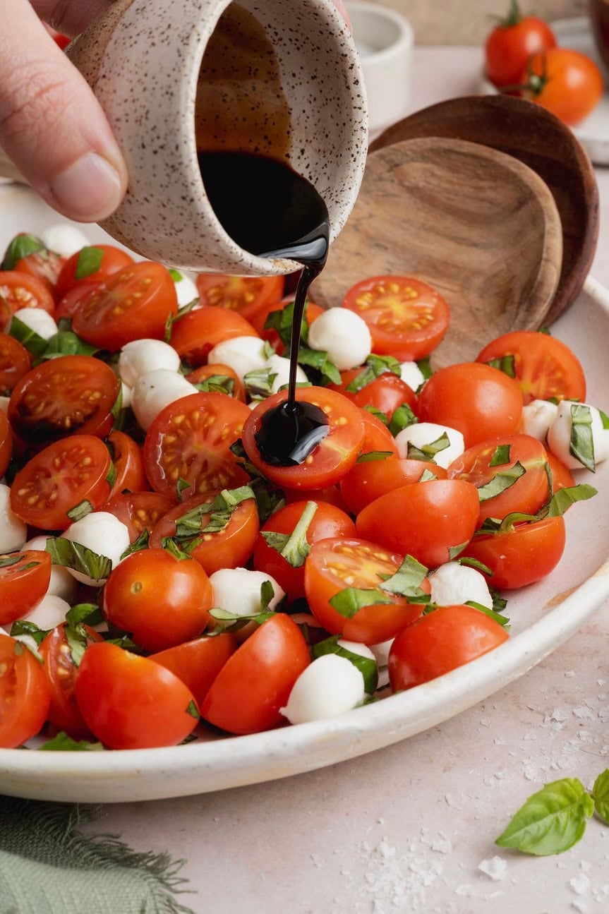 A hand drizzling balsamic glaze over a platter of caprese salad. 