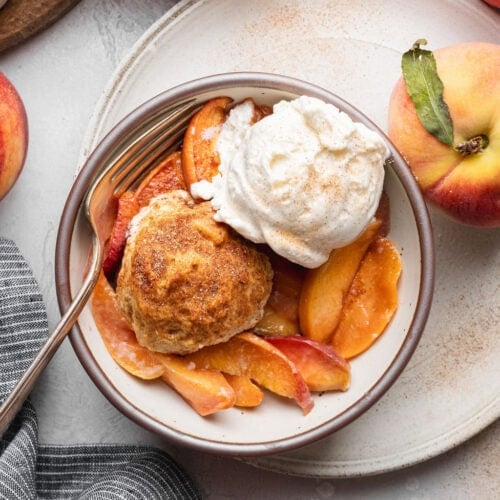 A bowl of peach cobbler topped with a biscuit and a scoop of vanilla ice cream, a fork on the side of the bowl.