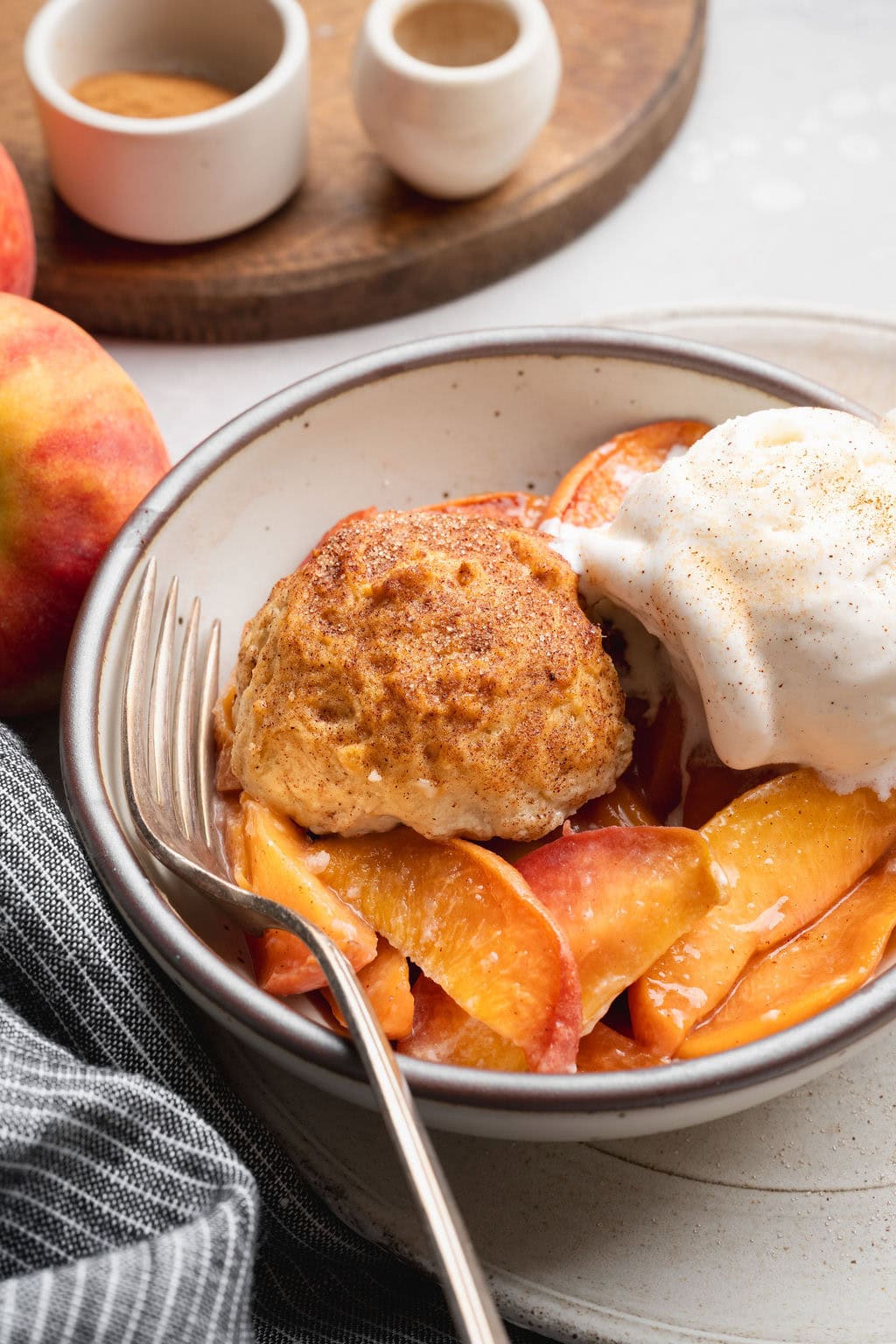 A bowl of homemade peach cobbler topped with a scoop of vanilla ice cream, a fork on the side.