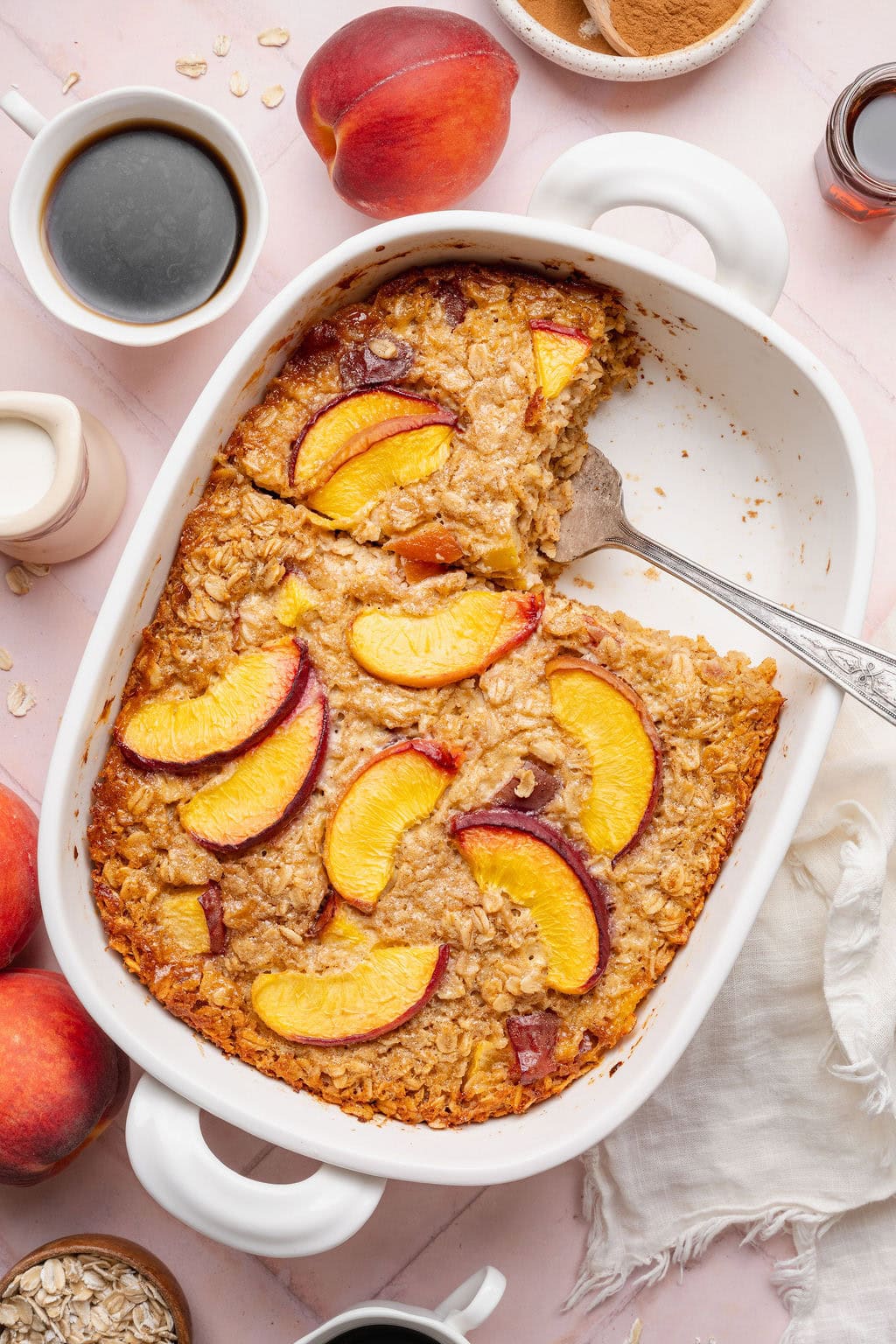 A baking dish of peach baked oatmeal with one square serving missing, a serving spoon sticking out under a slice.